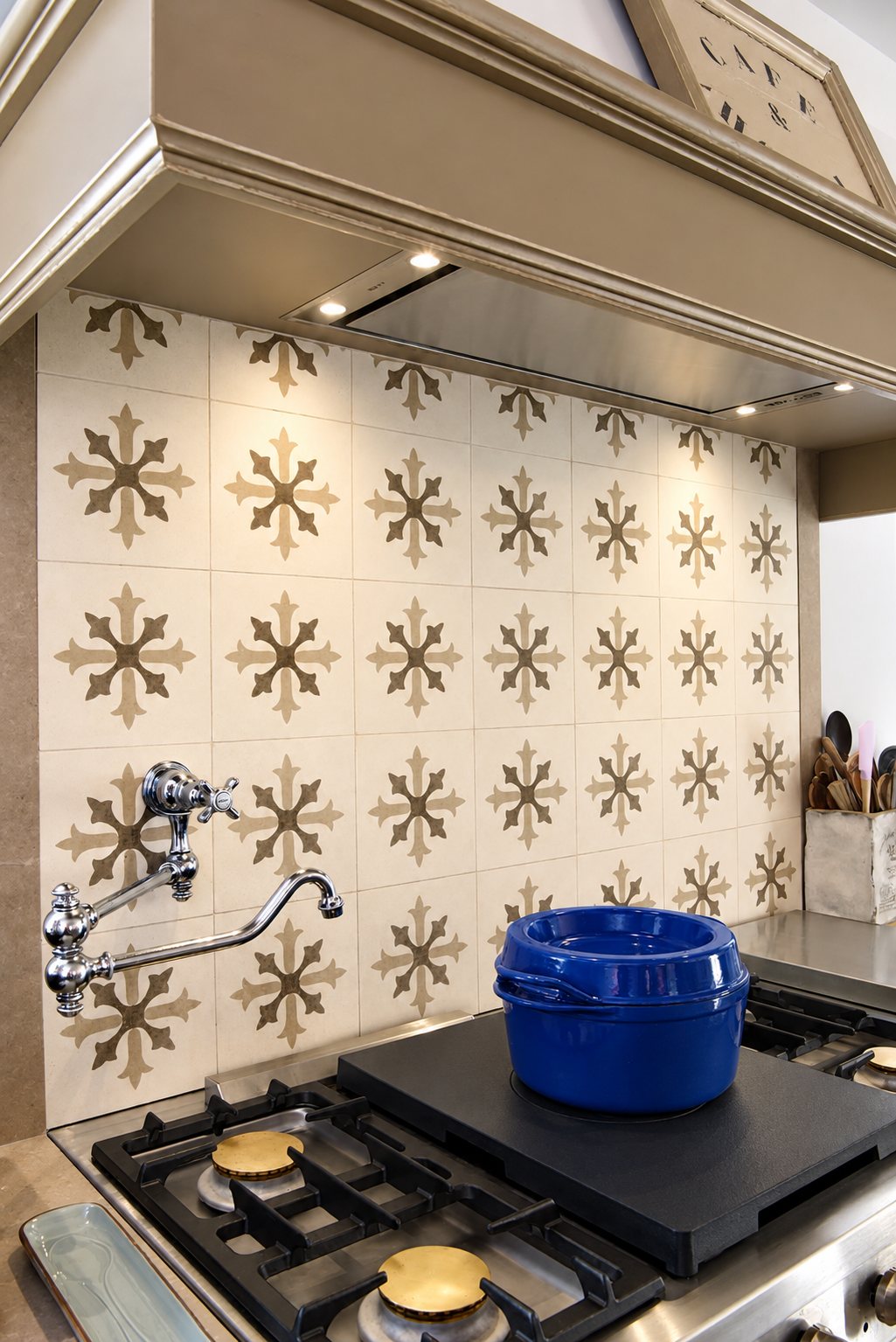 Une crédence de cuisine en carreaux beiges à motifs floraux surplombe une cuisinière à gaz, avec une cocotte bleue, un robinet chromé et une hotte en bois.