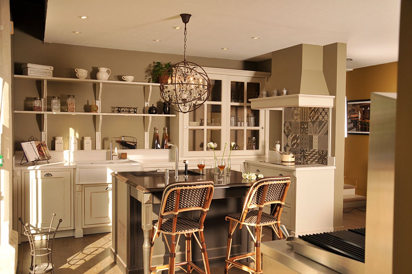 Showroom de cuisine de style classique aux tons beige et taupe, avec un îlot central, deux tabourets en rotin, un lustre à pampilles et une crédence en carreaux de ciment.