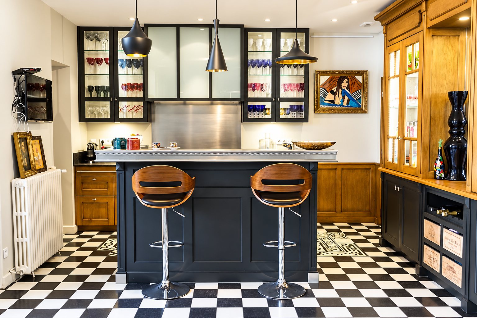 Cuisine au carrelage damier noir et blanc avec un îlot noir à plateau en zinc, deux tabourets en bois et des vitrines remplies de verres.