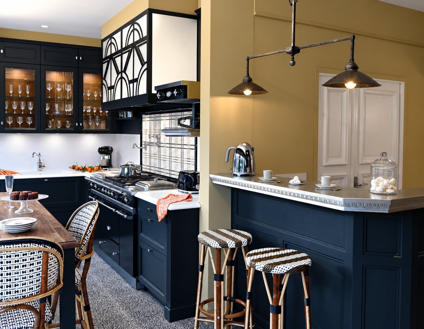 Cuisine aux meubles noirs et aux murs ocre, avec piano de cuisson, vitrines de verres, bar à plateau métallique et tabourets tressés.