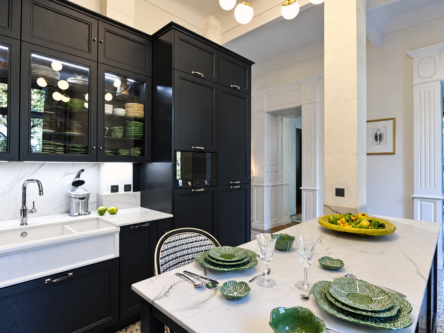 Cuisine de style ancien aux meubles noirs à vitrines, évier blanc et îlot en marbre dressé avec de la vaisselle verte en forme de feuilles de chou.