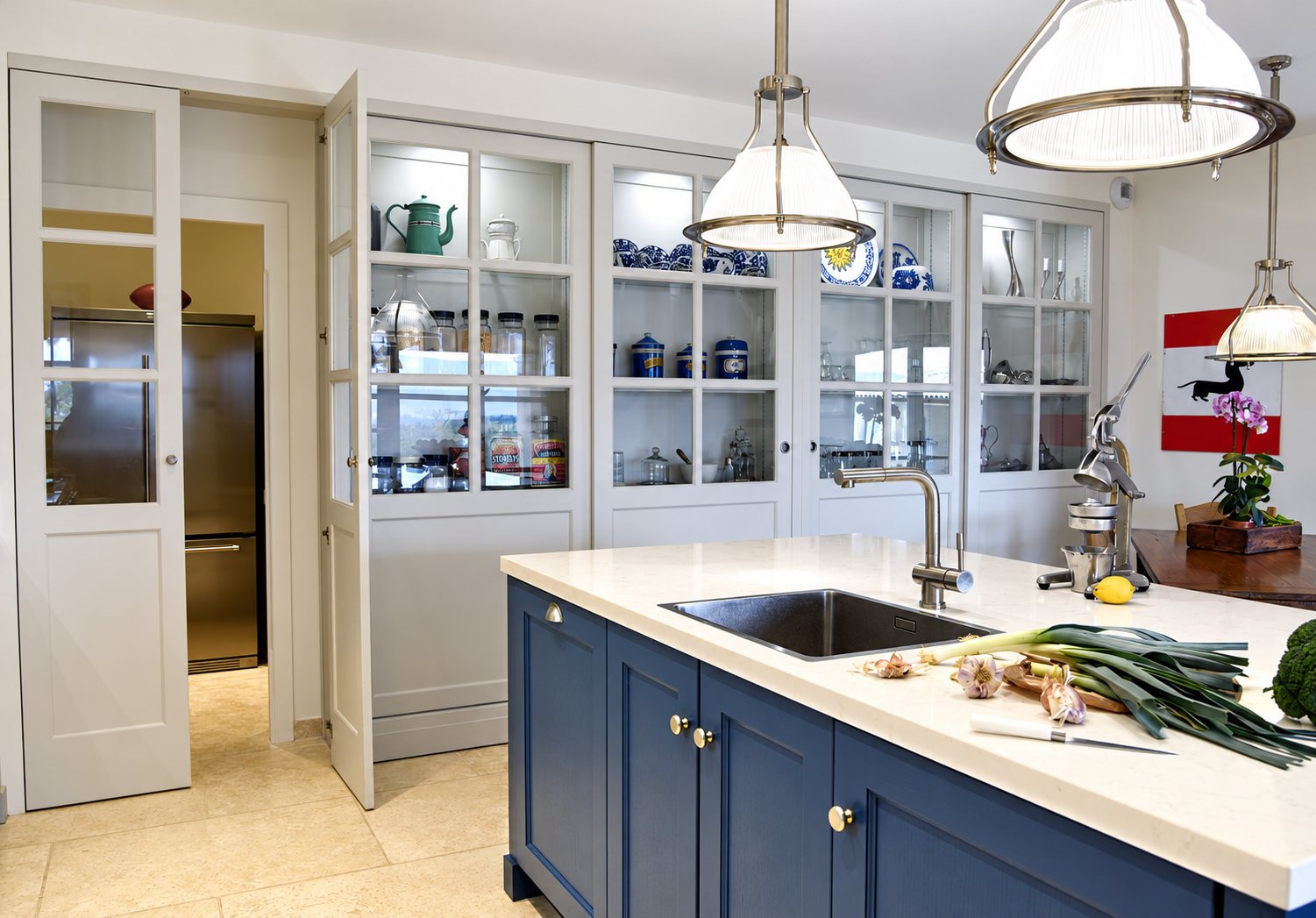 Cuisine lumineuse avec un îlot central bleu à plan de travail blanc, de grandes vitrines grises remplies de vaisselle et des suspensions en verre.