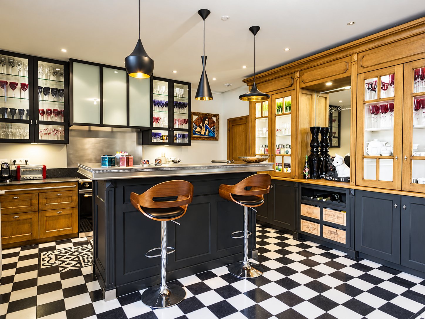 Cuisine aménagée avec un îlot central à plateau en zinc, deux tabourets en bois, des vitrines remplies de verres et un sol en damier noir et blanc.