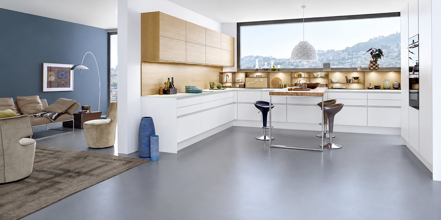 Grande cuisine moderne aux façades blanches et bois clair avec îlot et tabourets, ouverte sur un salon aux murs gris-bleu.