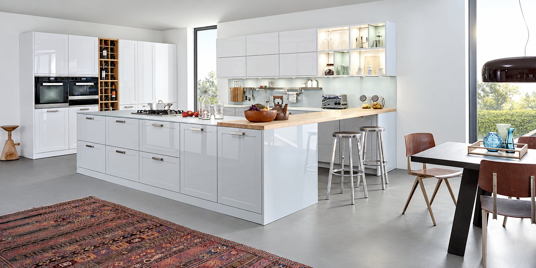 Cuisine moderne aux façades blanches brillantes avec un grand îlot, un plan de travail en bois, des tabourets en métal et une table à manger près d'une grande baie vitrée.