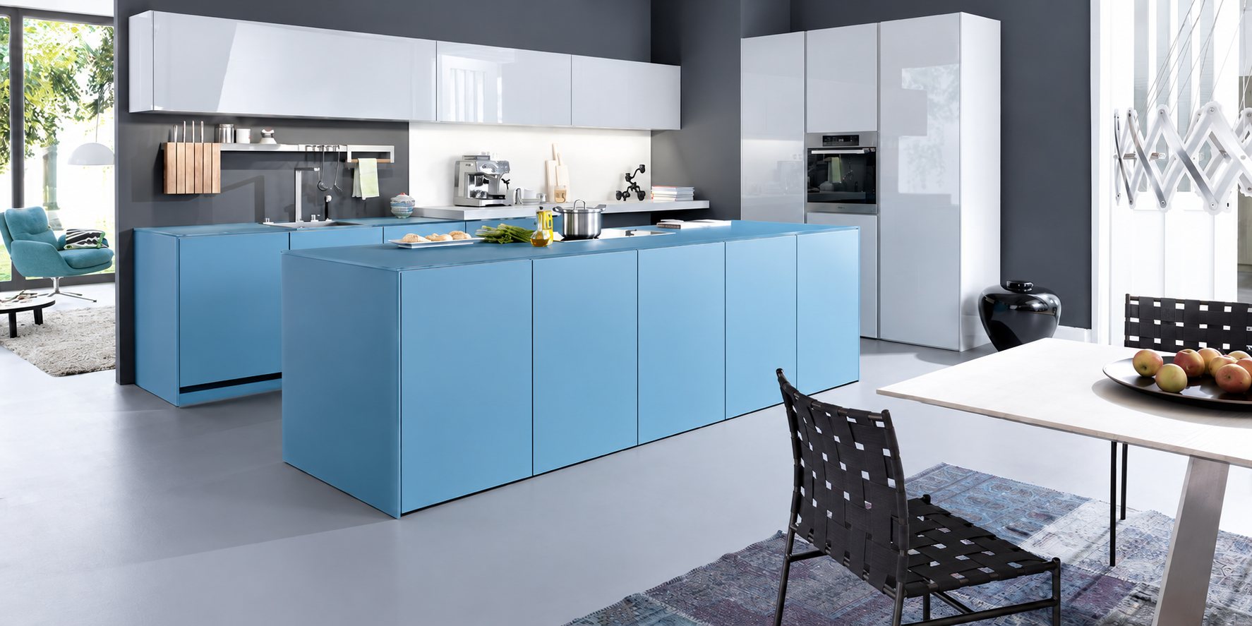 Cuisine contemporaine aux meubles bleu clair et gris laqué avec un grand îlot central, une table blanche, des chaises noires et un fauteuil bleu au fond.