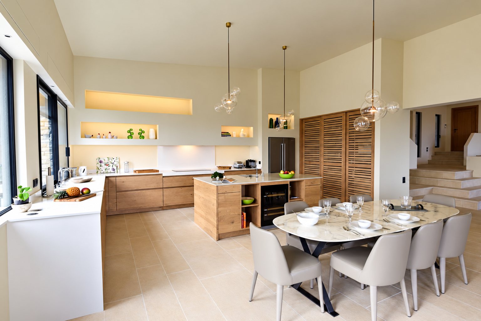 Grande cuisine ouverte aux façades en bois clair, avec îlot central, suspensions en verre et table ovale entourée de chaises beiges.