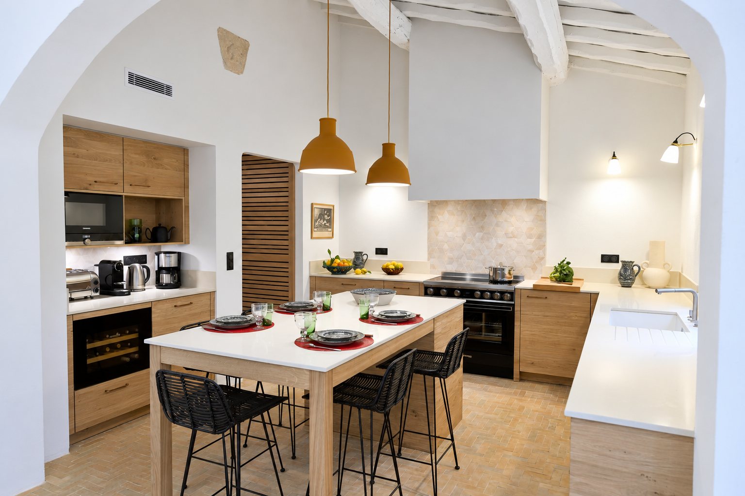 Cuisine lumineuse aux meubles en chêne clair, avec grand îlot dressé pour le repas, piano noir, suspensions ocre et poutres apparentes au plafond.