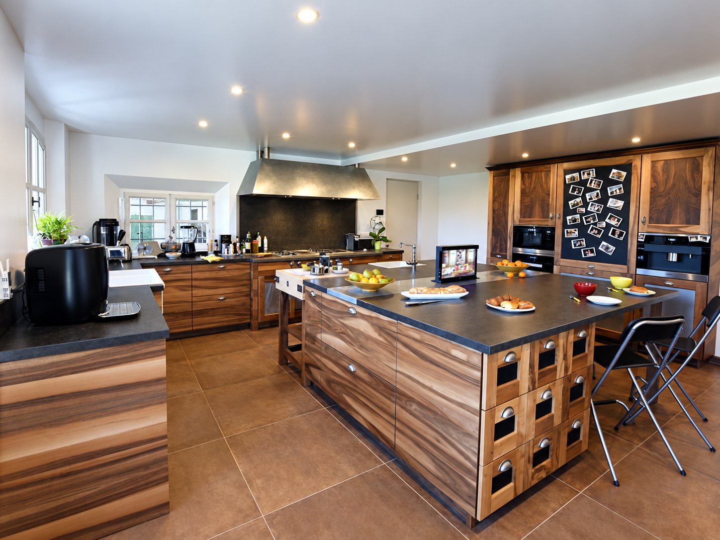 Cuisine spacieuse aux meubles en noyer, avec grand îlot central à plan sombre, hotte en inox et carrelage brun au sol.