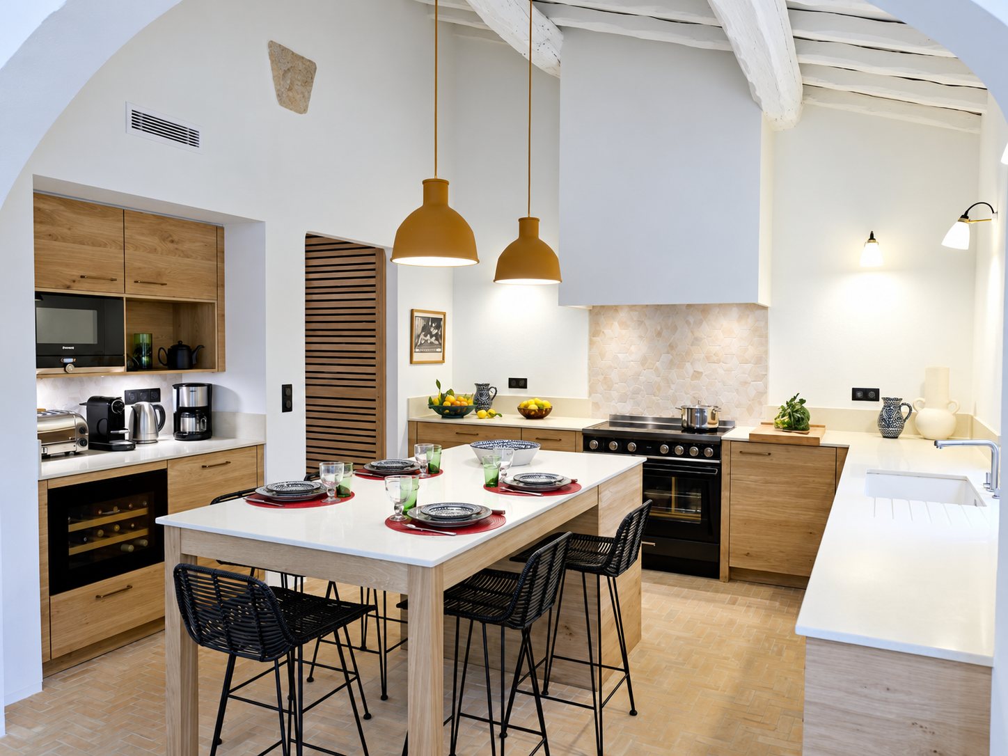 Cuisine lumineuse aux meubles en chêne clair avec un îlot dressé pour un repas, des tabourets noirs, deux suspensions ocre et des poutres blanches apparentes.