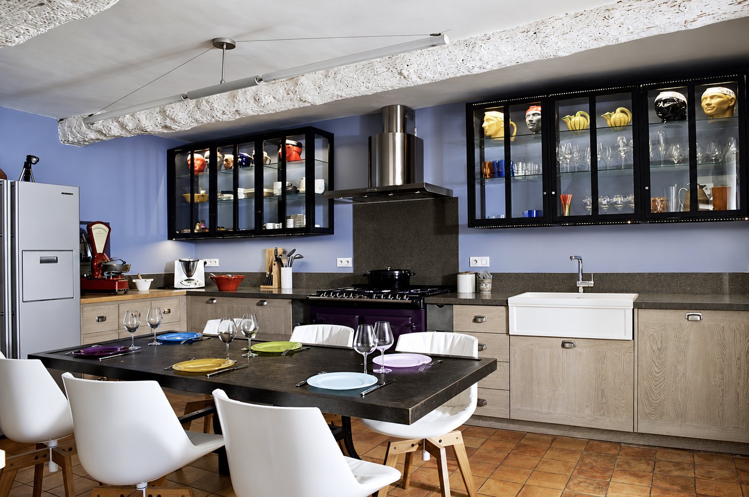 Cuisine aux murs bleu-violet avec meubles en chêne clair, vitrines noires, évier en céramique et table noire dressée entourée de chaises blanches.
