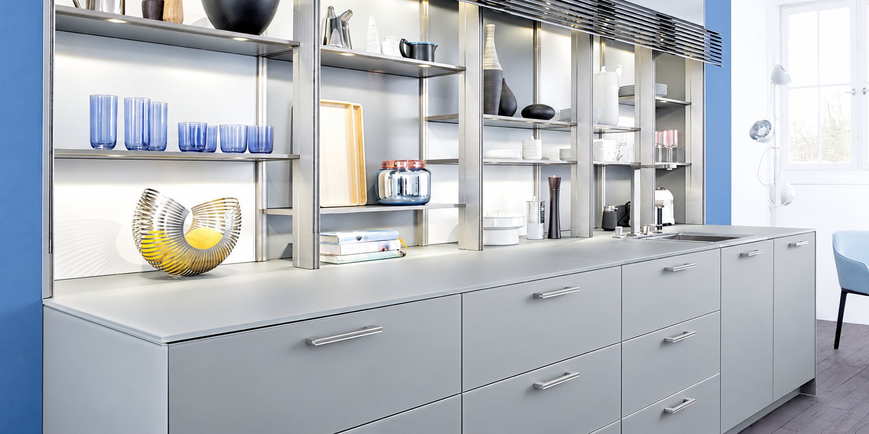 Cuisine contemporaine aux façades gris clair, surmontée d'étagères métalliques garnies de verres bleus, de vaisselle et d'objets décoratifs, avec des murs bleus de chaque côté.