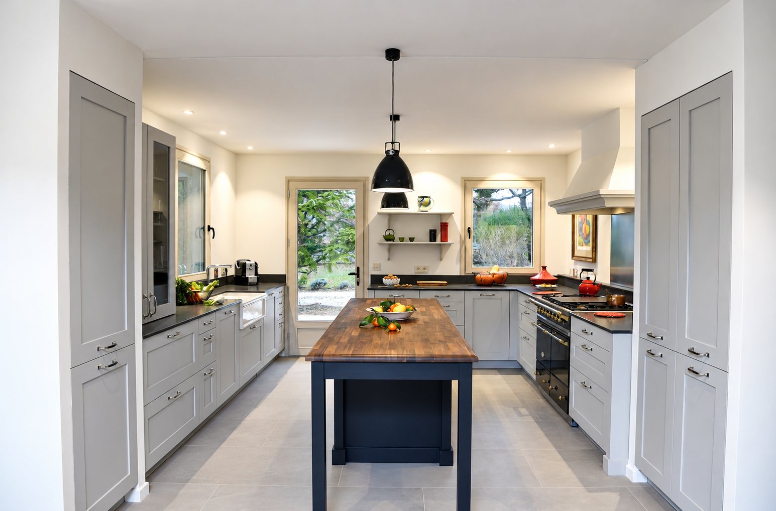 Cuisine équipée aux meubles gris clair avec un îlot central bleu foncé au plateau en bois, deux suspensions noires et de larges fenêtres donnant sur le jardin.