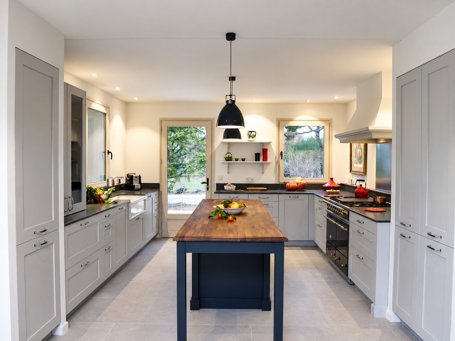 Cuisine équipée aux façades gris clair avec un îlot central bleu foncé et plateau en bois, deux suspensions noires et de grandes fenêtres donnant sur le jardin.