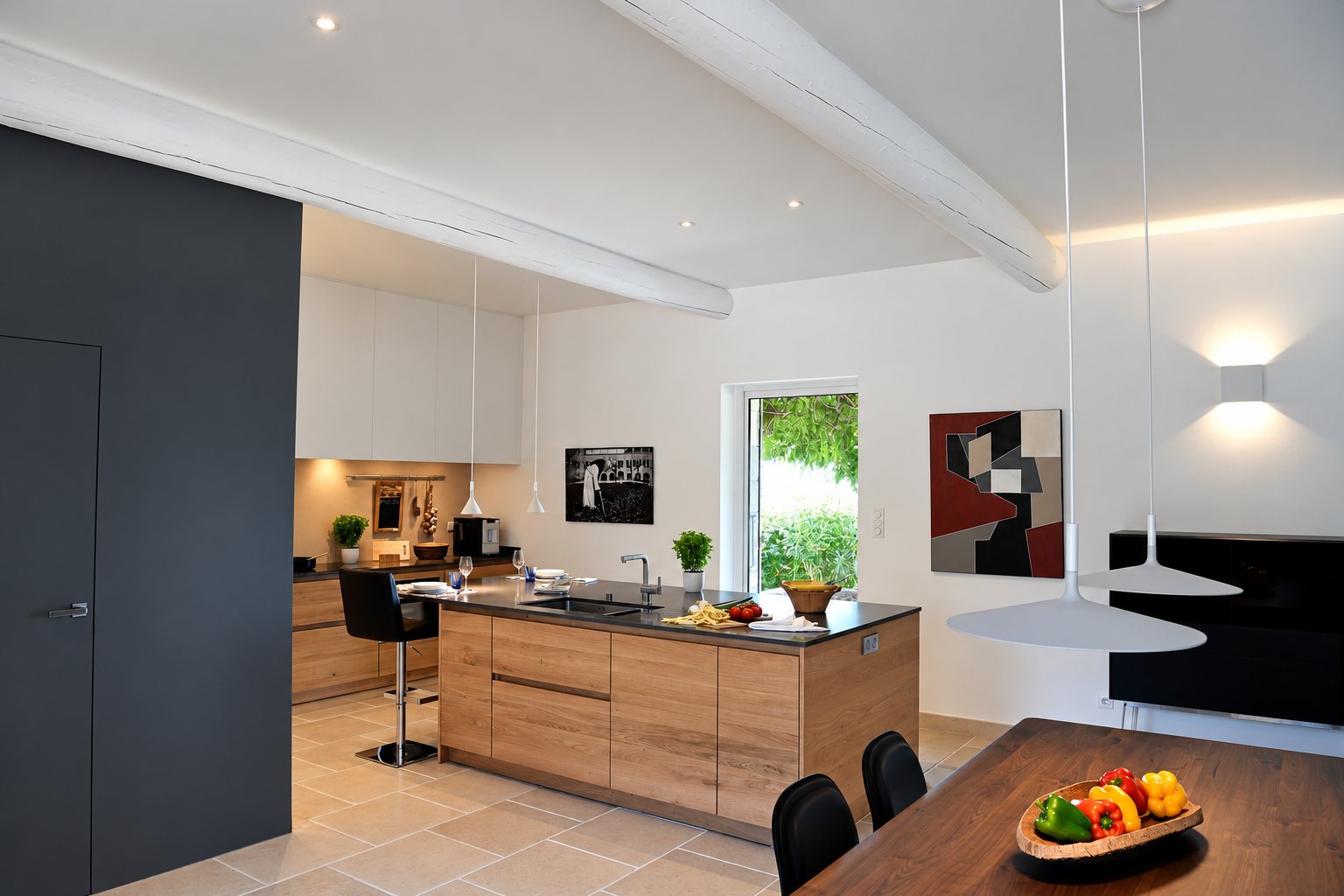 Cuisine contemporaine lumineuse avec îlot central en bois clair, plan de travail sombre, poutres blanches apparentes et table avec corbeille de poivrons.