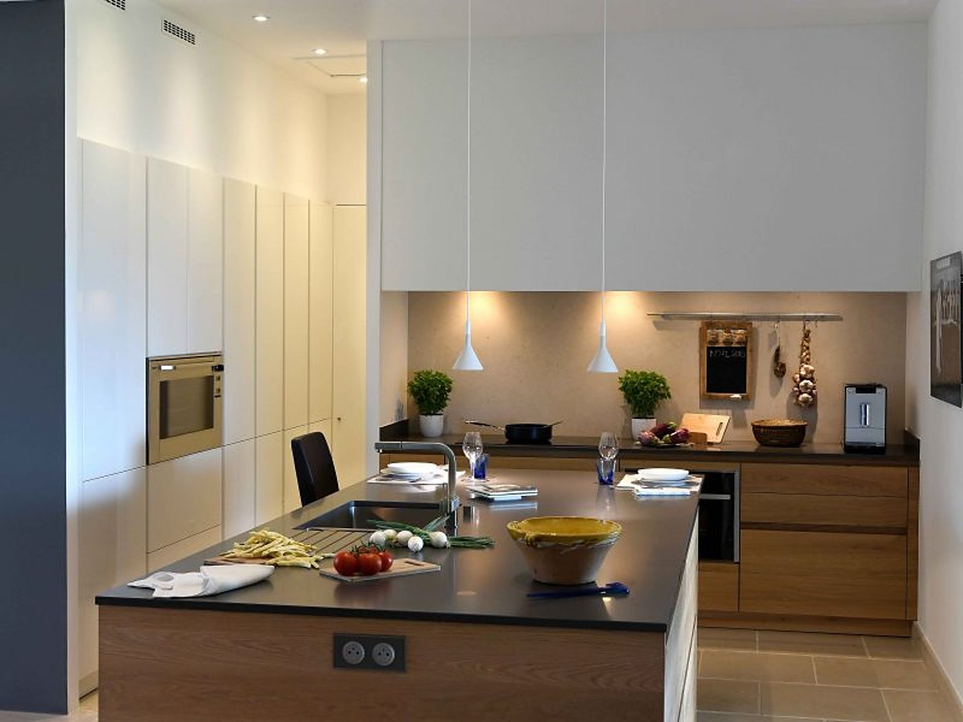 Cuisine contemporaine aux façades blanches et en bois avec un îlot central à plan de travail sombre, des suspensions et des légumes frais préparés.