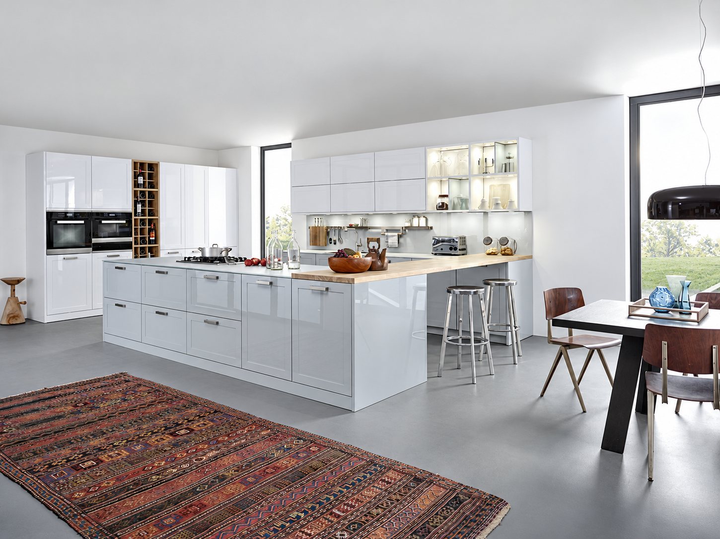 Cuisine contemporaine aux façades laquées blanches et grises, avec un grand îlot à plan de travail en bois, des tabourets, une table à manger et un tapis coloré.