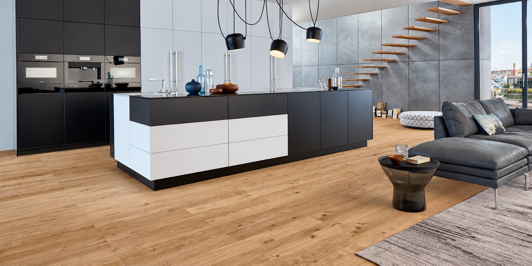 Cuisine contemporaine noire et blanche avec grand îlot central, suspensions noires, parquet clair et salon avec canapé gris et escalier suspendu.