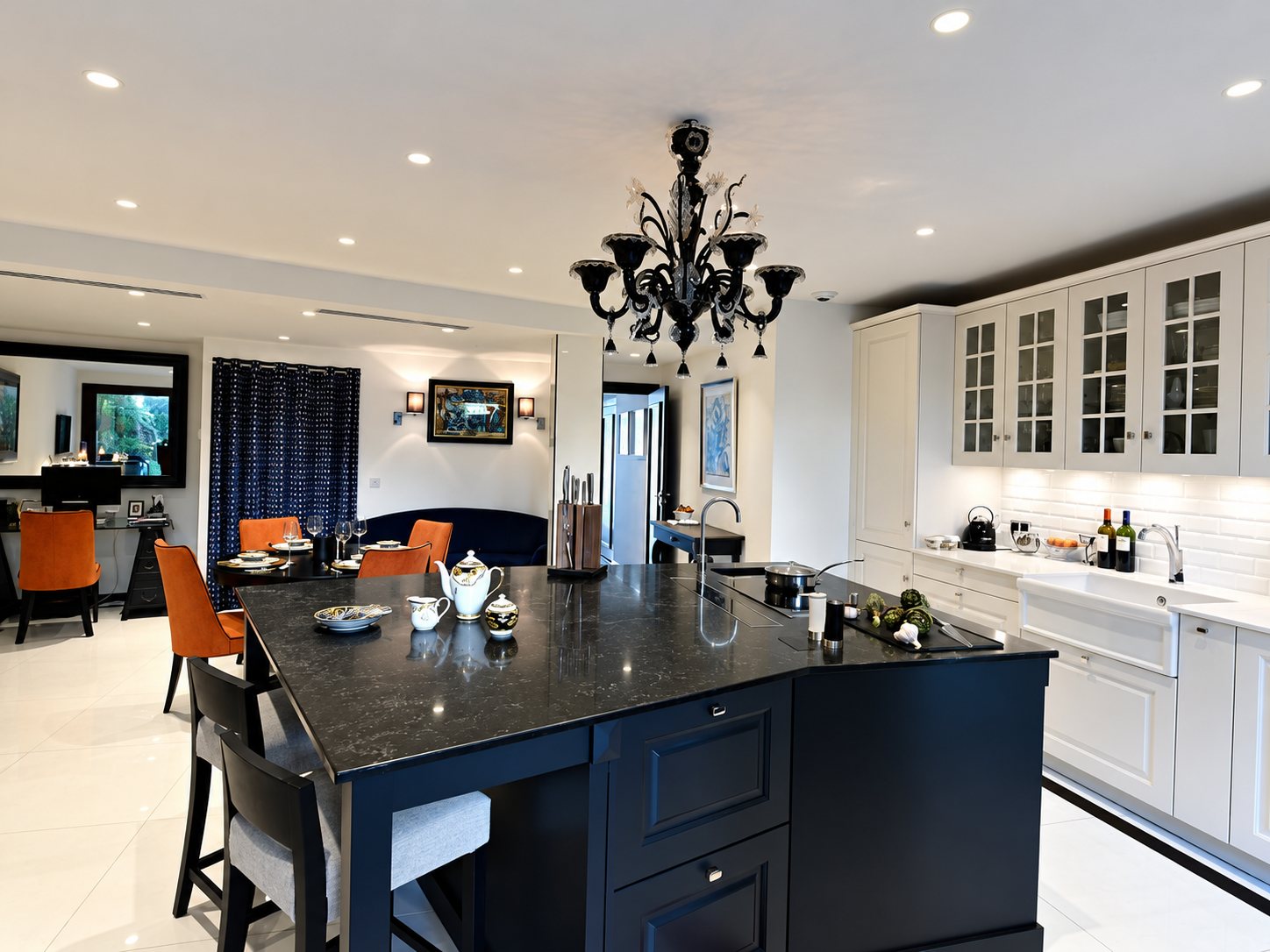 Cuisine ouverte avec îlot central noir, placards blancs, lustre noir et coin repas aux chaises orange.