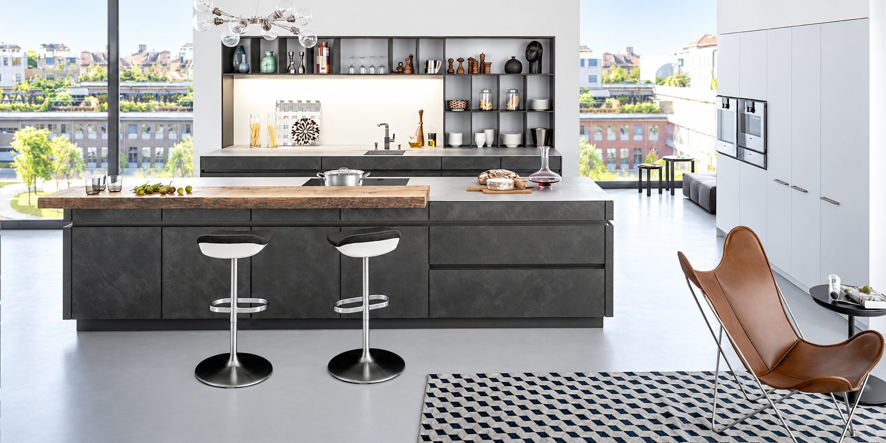 Cuisine contemporaine lumineuse avec un grand îlot gris anthracite, un plan de bar en bois brut, deux tabourets et un fauteuil en cuir marron au premier plan.