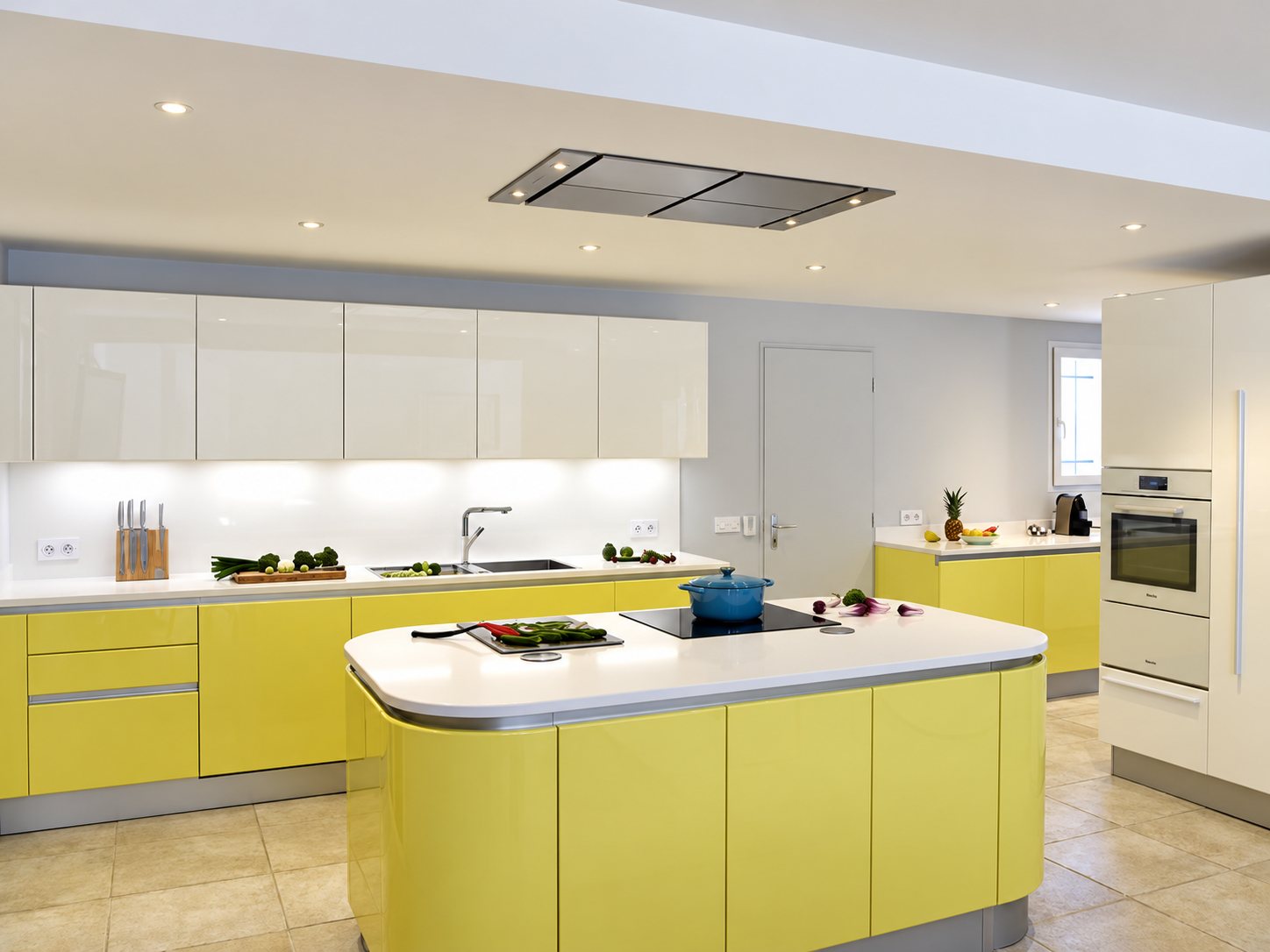 Cuisine équipée moderne aux façades jaunes et blanches laquées, avec un îlot central arrondi, une plaque de cuisson et une hotte plafonnière.