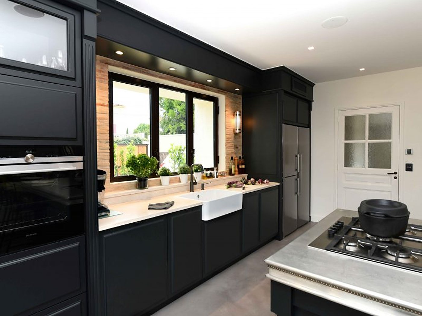 Cuisine contemporaine aux meubles noirs, avec évier en céramique blanc sous une fenêtre, mur de briques, four encastré et îlot avec plaque de cuisson.