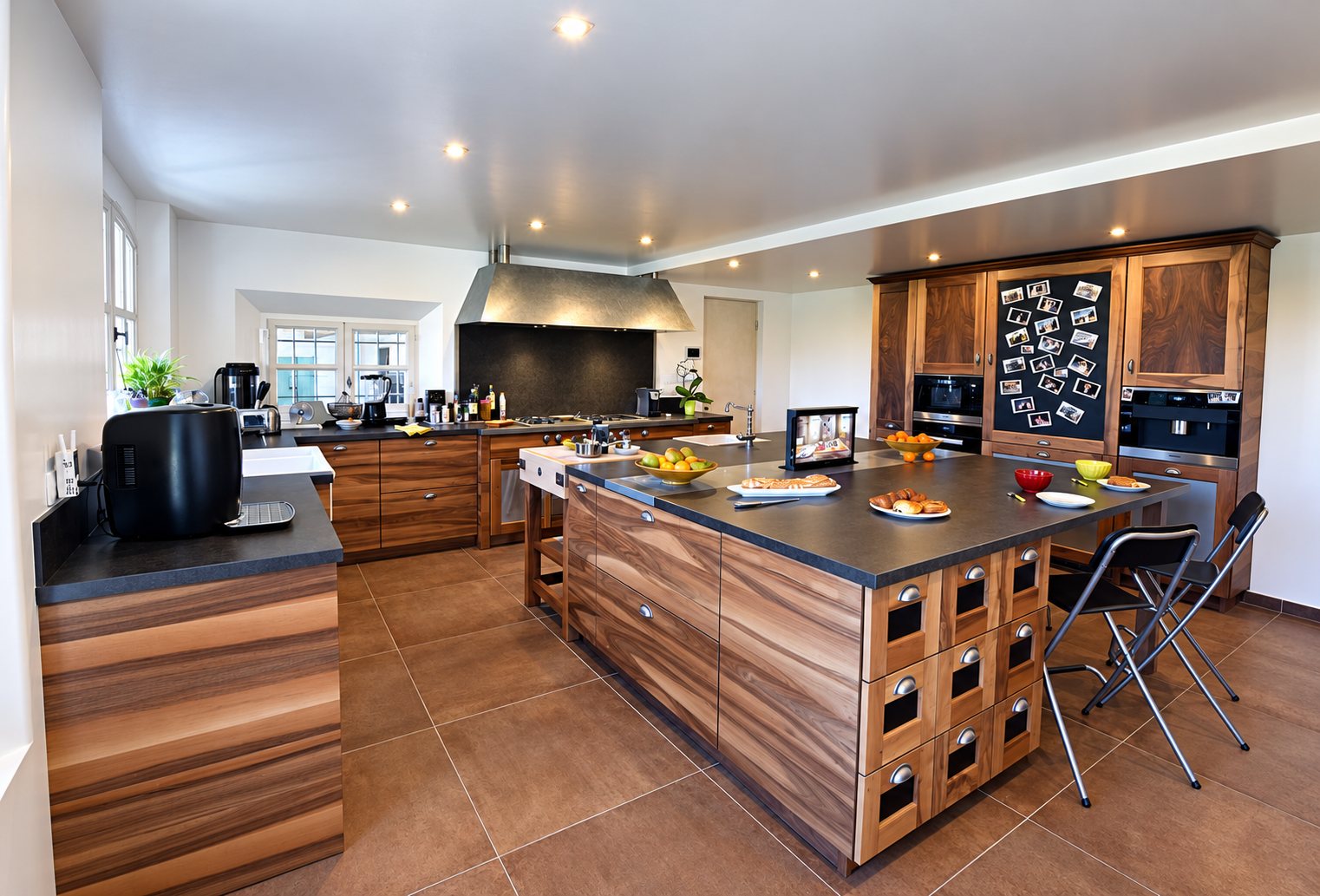 Vaste cuisine contemporaine aux meubles en noyer, dotée d'un grand îlot central à plan de travail sombre, d'une hotte inox et d'un carrelage brun.