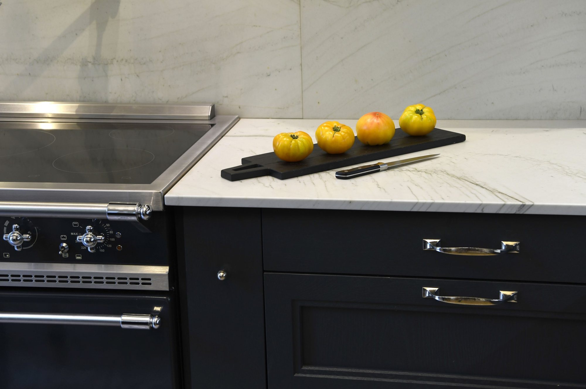 Angle de cuisine avec piano de cuisson noir et inox, meubles anthracite, plan de travail en marbre clair et quatre tomates jaunes sur une planche en bois noir avec un couteau.