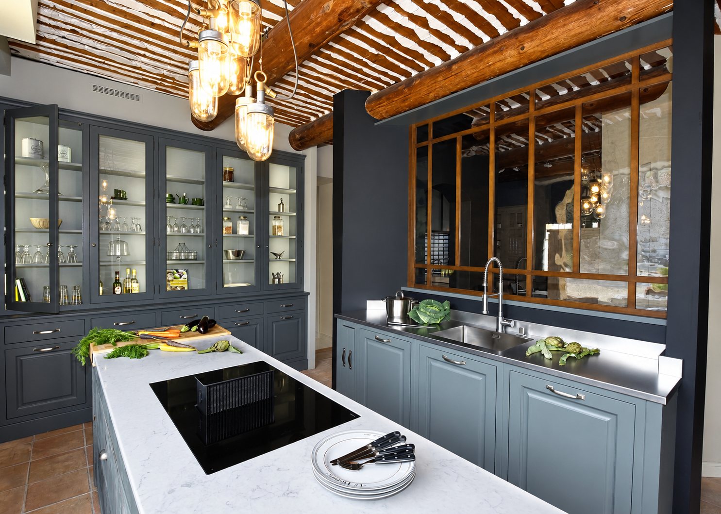 Cuisine aux meubles gris-bleu avec grande vitrine, îlot à plaque de cuisson, évier en inox et plafond à poutres en bois rondes.