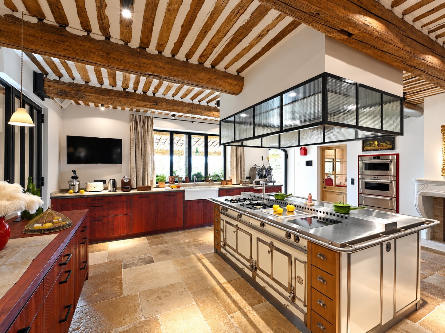 Grande cuisine rustique au plafond à poutres de bois, dotée d'un îlot central en inox avec piano de cuisson, de meubles rouge foncé et d'un sol en pierre claire.