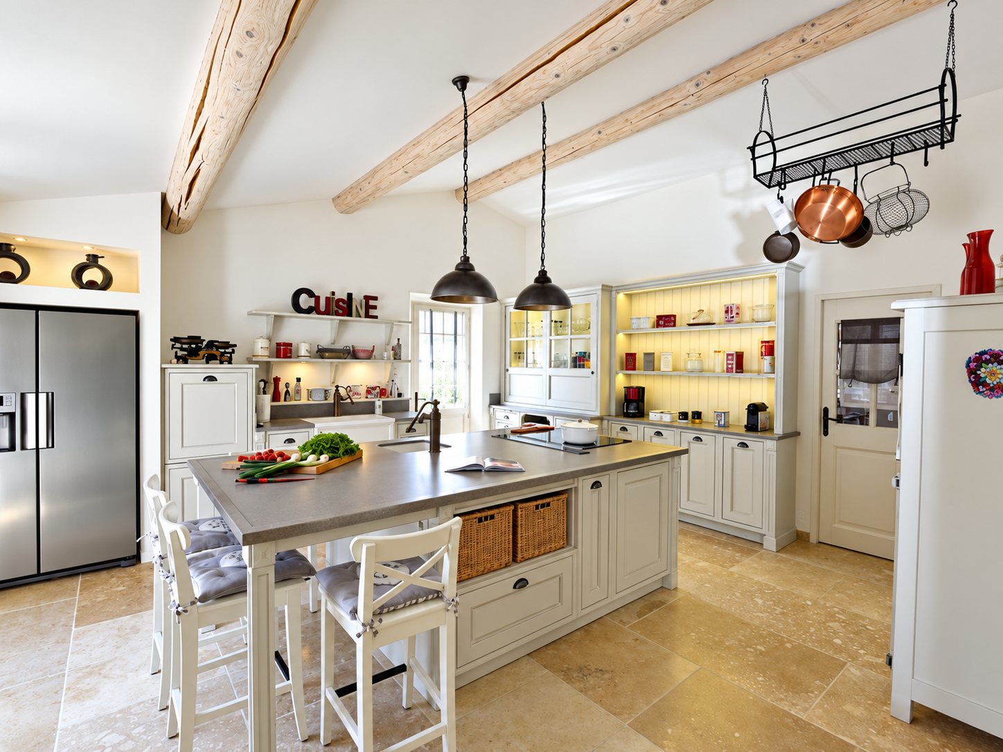 Vaste cuisine de style provençal avec îlot central en inox, tabourets blancs, poutres claires au plafond, buffet à fond jaune et sol en dalles de pierre.