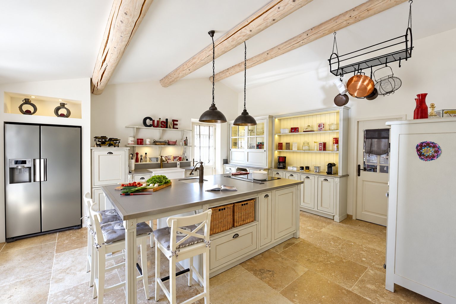 Une cuisine de style provençal avec un grand îlot central, des meubles gris clair, des poutres blanchies, un réfrigérateur américain et un sol en pierre claire.