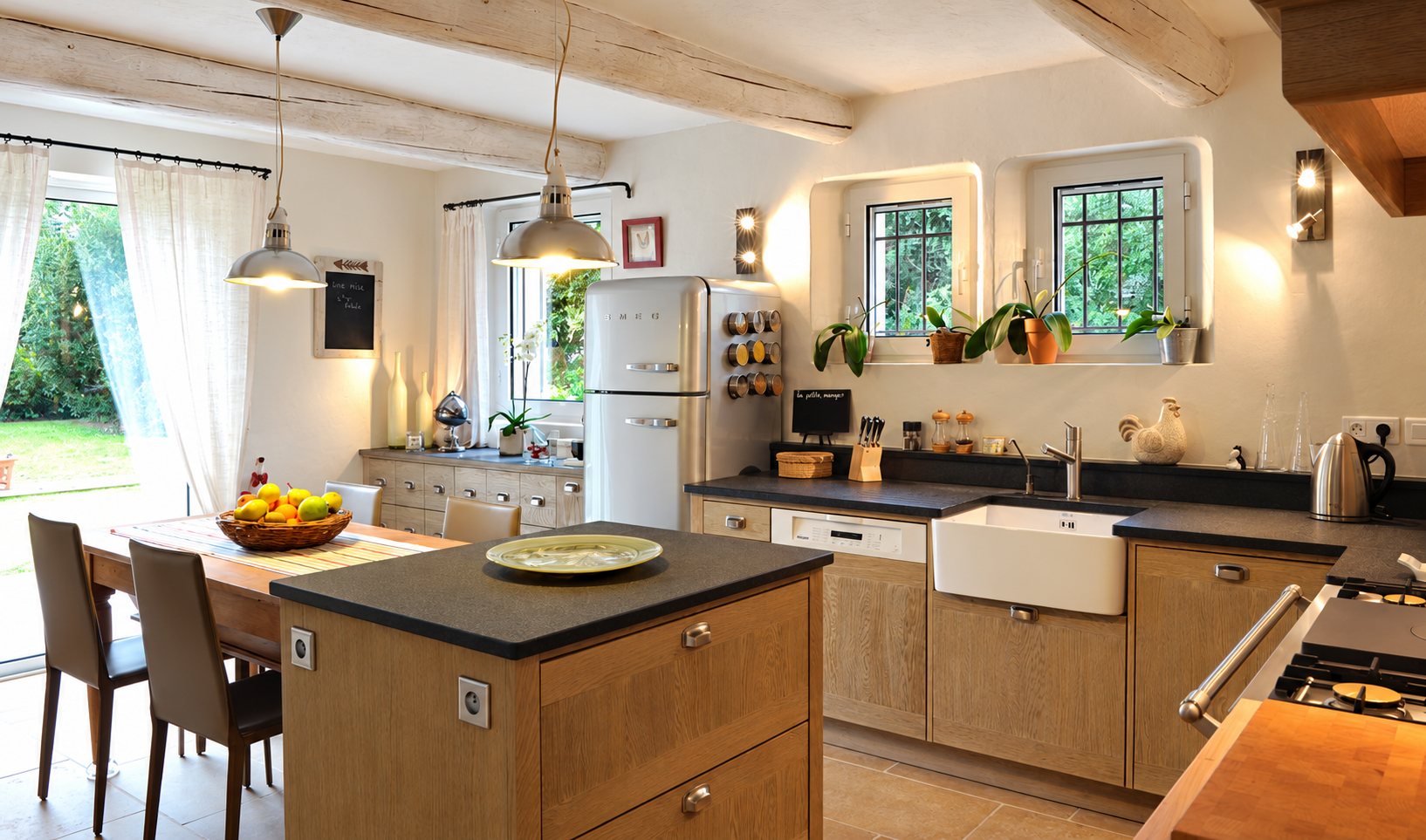 Cuisine rustique et lumineuse avec îlot en bois, plan de travail sombre, évier en céramique blanche, réfrigérateur rétro et table près d'une porte-fenêtre donnant sur le jardin.