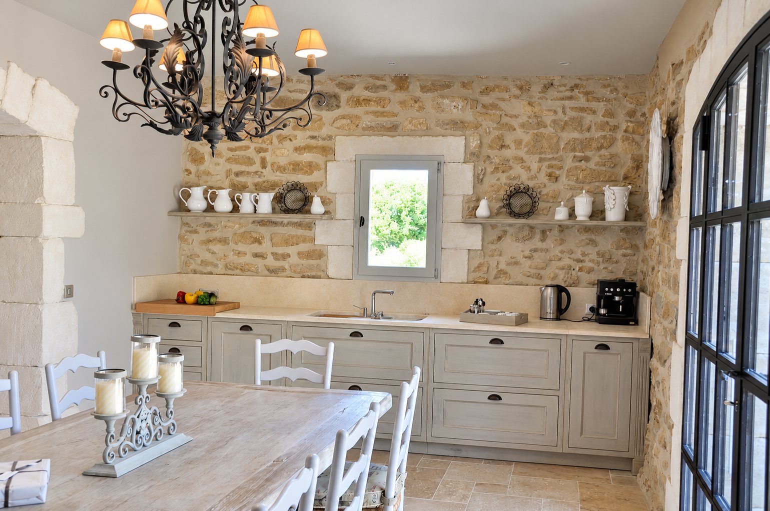 Cuisine rustique aux murs de pierre apparente, avec meubles gris clair, lustre en fer forgé et grande table en bois entourée de chaises blanches.
