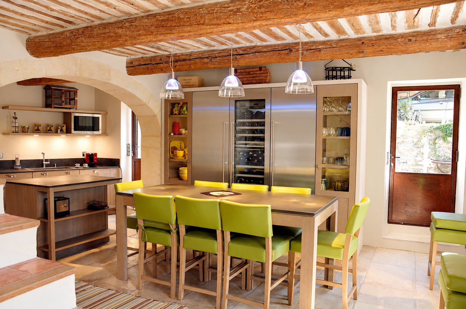 Cuisine rustique aux poutres apparentes avec une table haute en bois entourée de chaises vertes, un îlot et des armoires en inox et vitrées.
