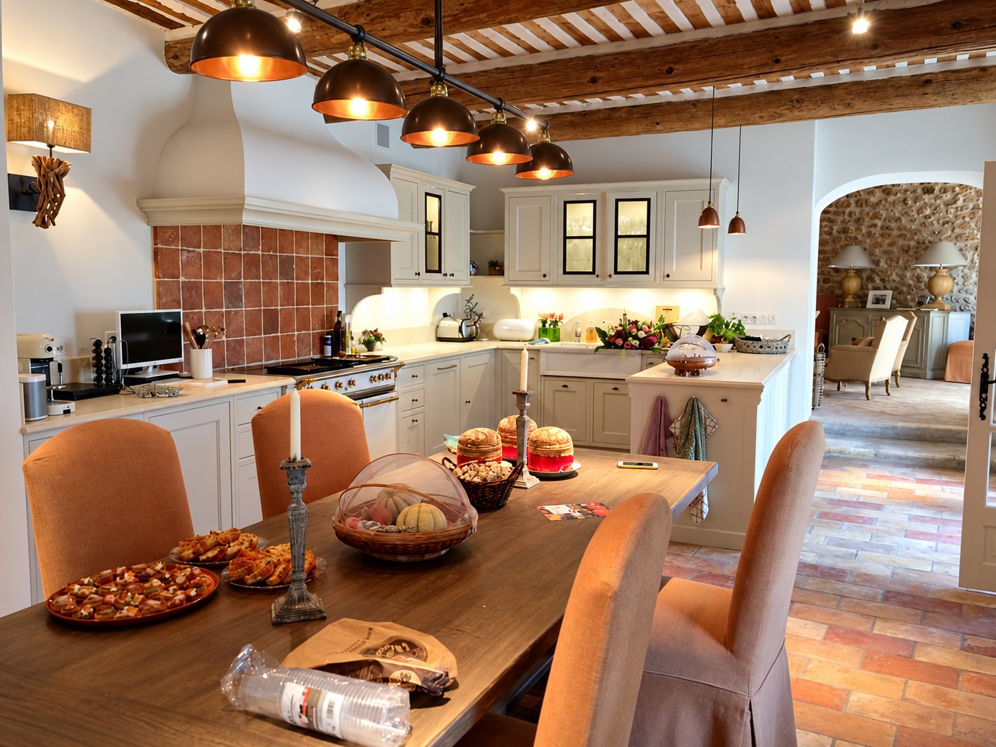 Grande cuisine provençale aux poutres apparentes et au sol en terre cuite, avec une longue table en bois garnie de pains et de plats salés.