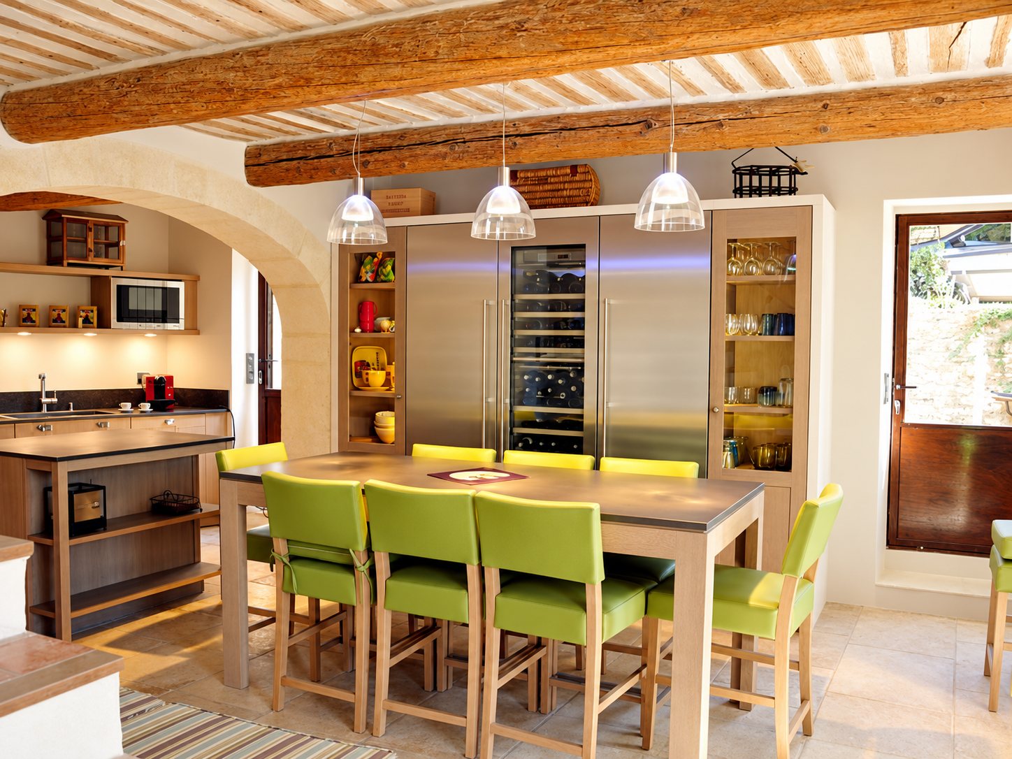 Cuisine rustique aux poutres apparentes, équipée d'une grande table haute en bois entourée de chaises vertes et d'un cellier à vin intégré dans une armoire en inox.