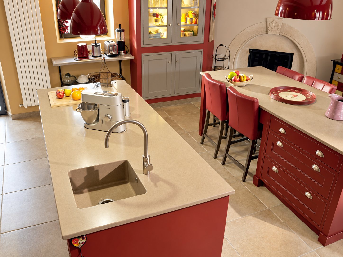 Cuisine aux meubles rouge bordeaux avec un îlot central à plan de travail beige, évier intégré, robinet moderne et tabourets rouges.