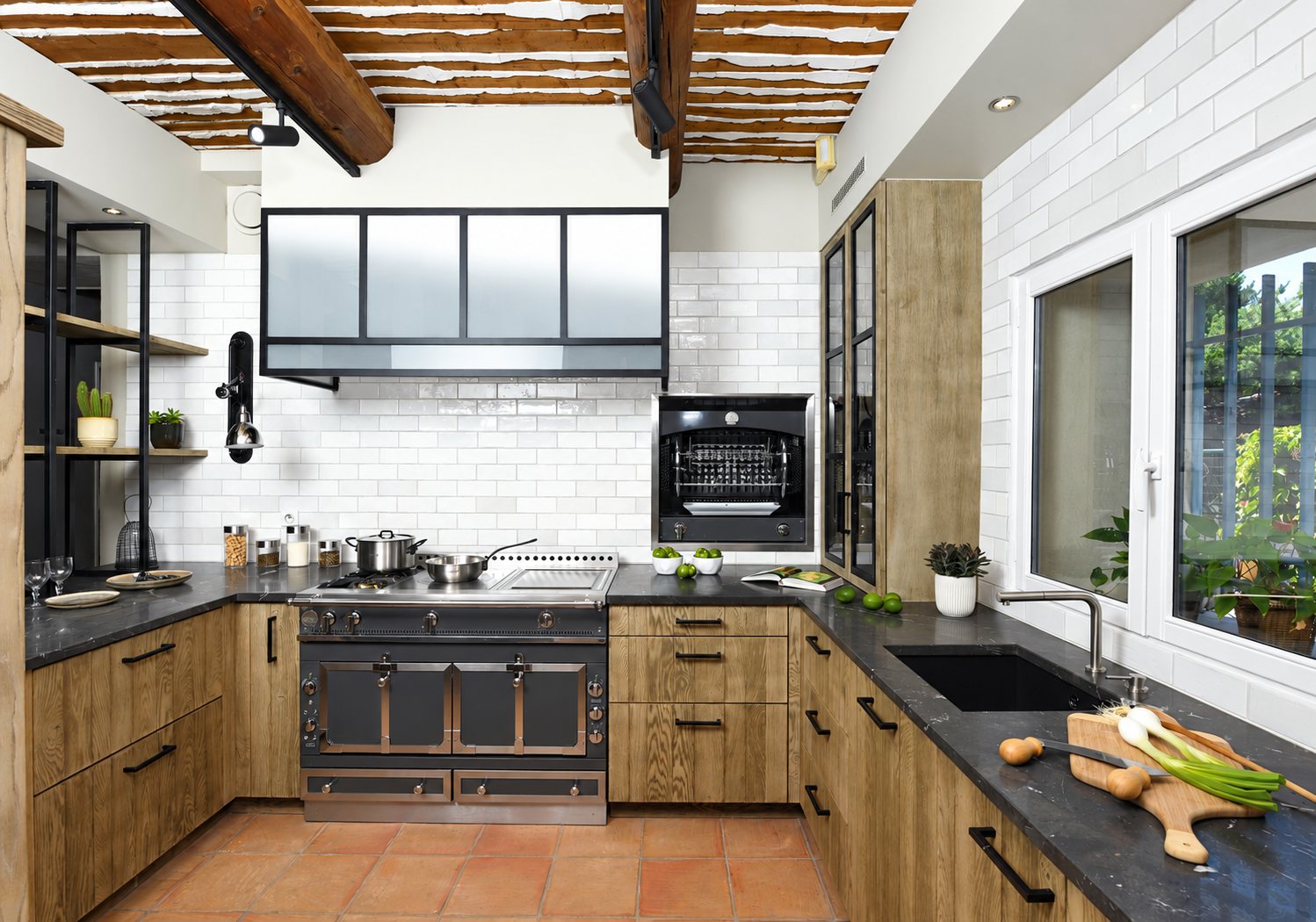 Cuisine aménagée avec meubles en chêne, plan de travail en pierre noire, piano de cuisson anthracite, four encastré et poutres apparentes au plafond.