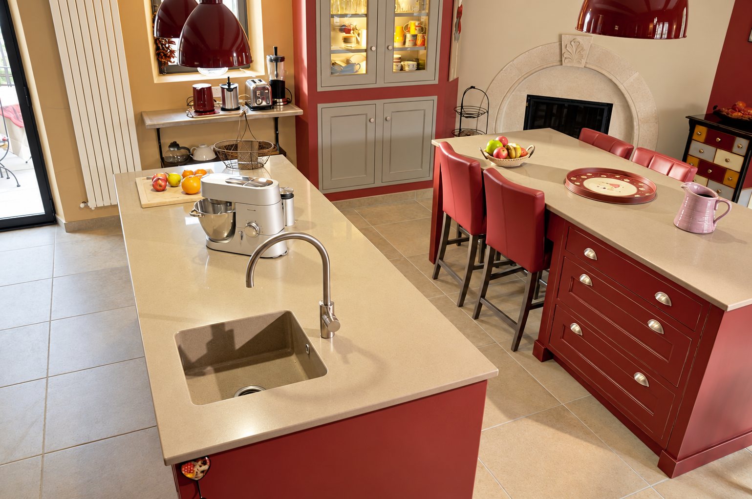 Cuisine aménagée avec deux îlots rouges à plan de travail beige, évier avec robinet chromé, tabourets en cuir rouge et cheminée en pierre.