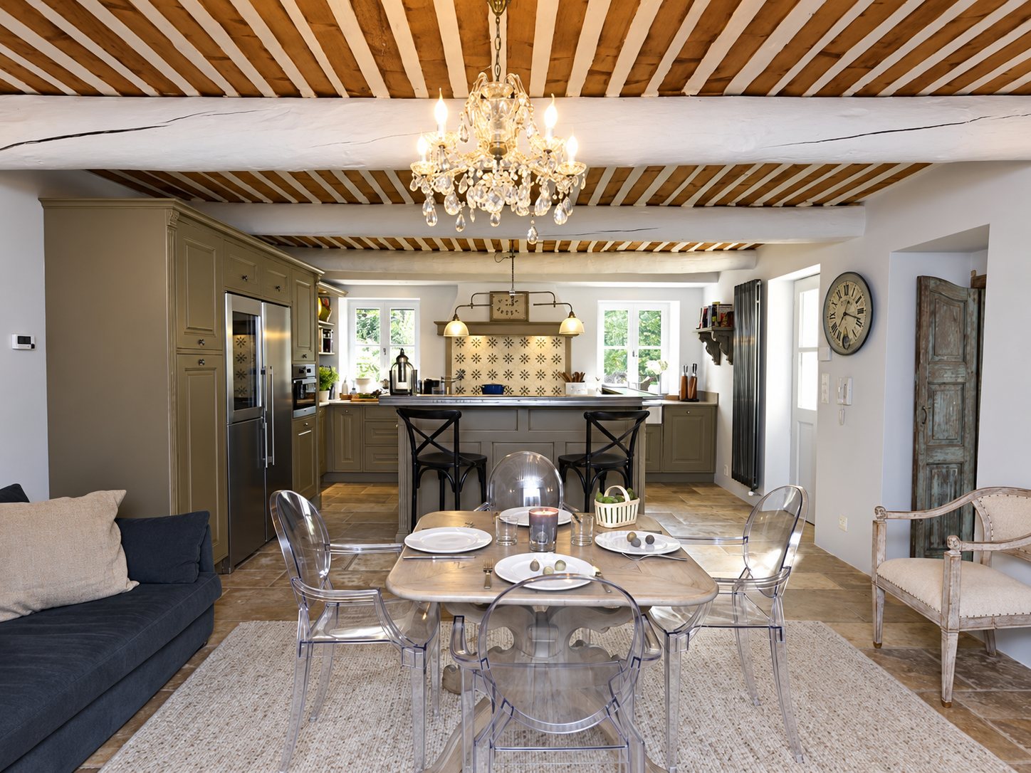 Pièce de vie provençale avec cuisine ouverte grise, table dressée entourée de chaises transparentes, lustre à pampilles et plafond en lattes de bois.