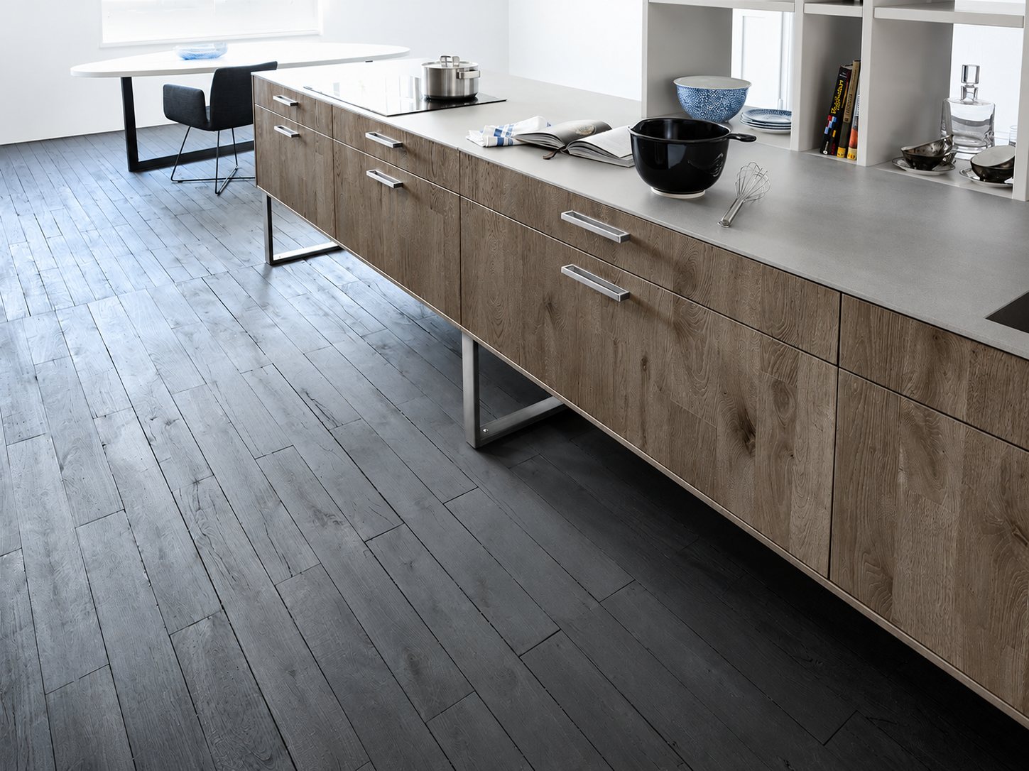Un îlot de cuisine moderne aux façades en chêne vieilli et plan de travail gris, posé sur un parquet gris foncé, avec une table ovale en arrière-plan.