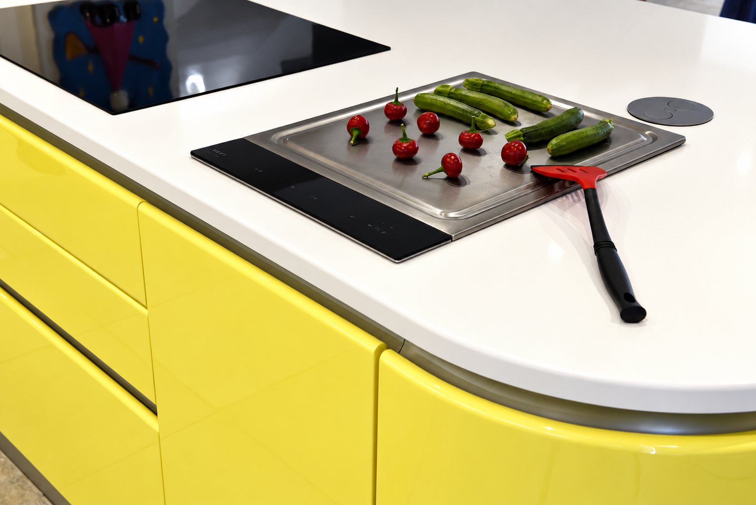 Îlot de cuisine aux façades jaune brillant et plan de travail blanc arrondi, avec une plaque de cuisson posée de courgettes, de petits piments rouges et une spatule rouge.