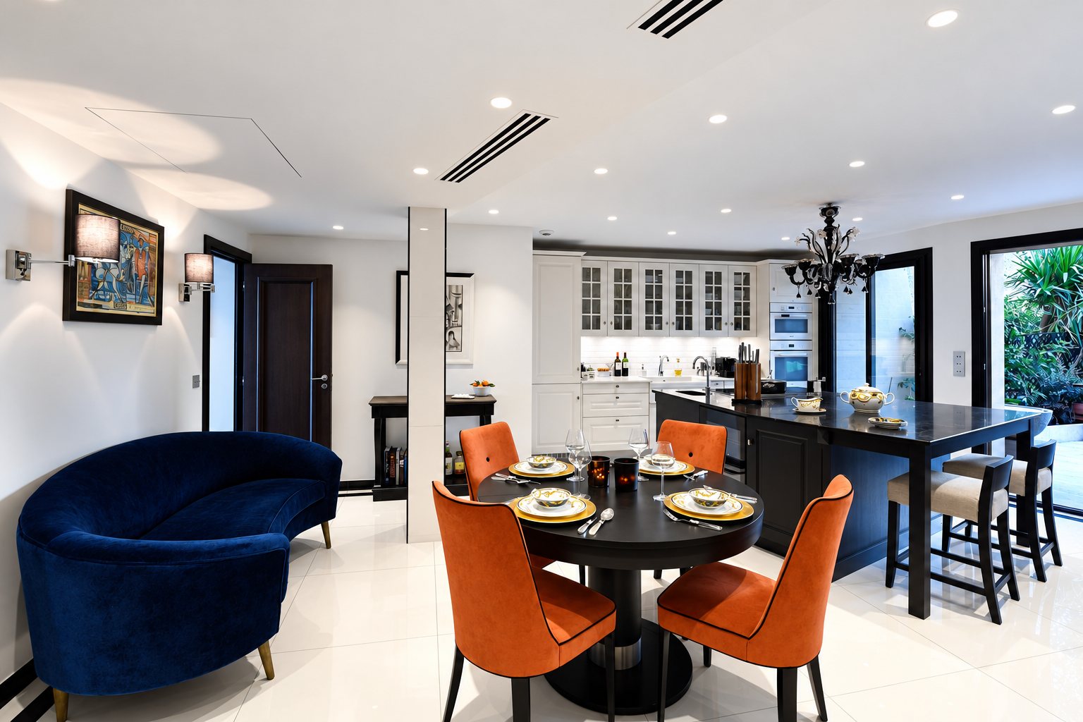 Intérieur lumineux d'un appartement avec canapé en velours bleu, table ronde dressée entourée de fauteuils orange et cuisine ouverte avec îlot noir.